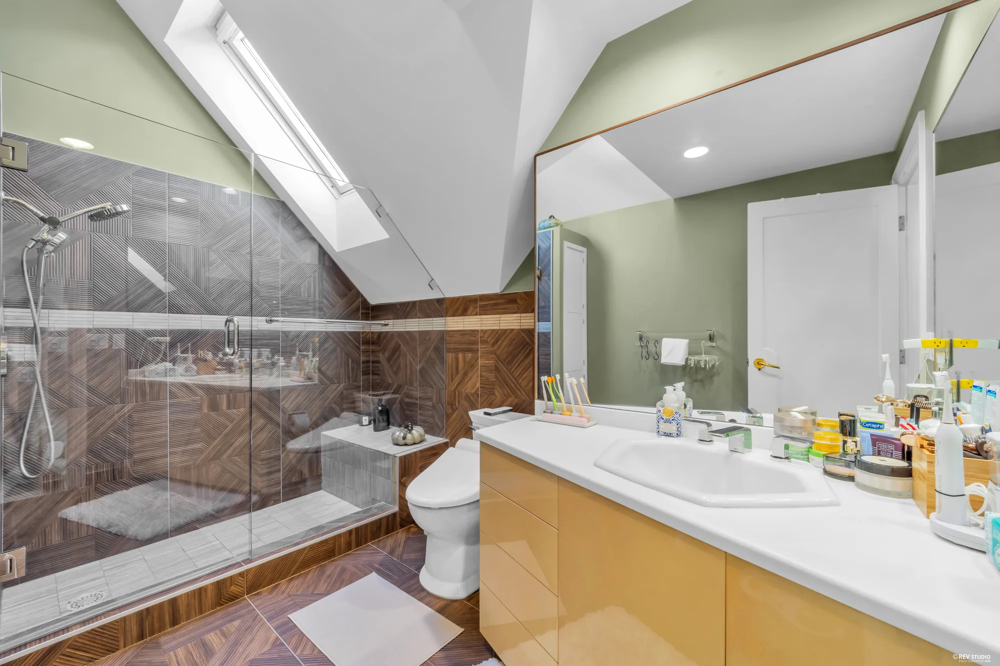 The upper bathroom: a glass shower in wood-look tile, a skylight, a vanity under a wide mirror