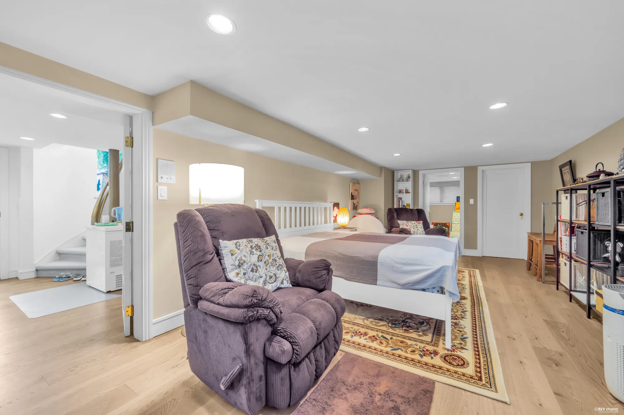 The lower bedroom: a bed and a recliner on new hardwood, a door to the hall on the left, a low ceiling with pot lights