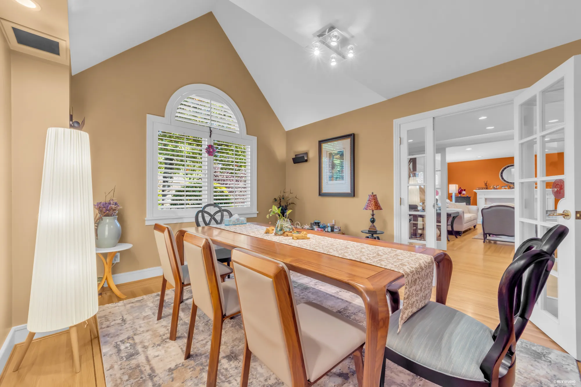 The dining room: a long wooden table under a vaulted ceiling, an arched window over shutters, French doors to the family room on the right