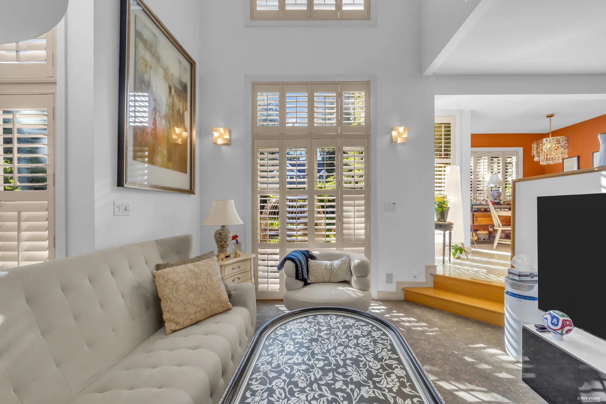 The family room: cream sofas around a brass-edged table, plantation shutters two storeys high, the steps up to the kitchen on the right