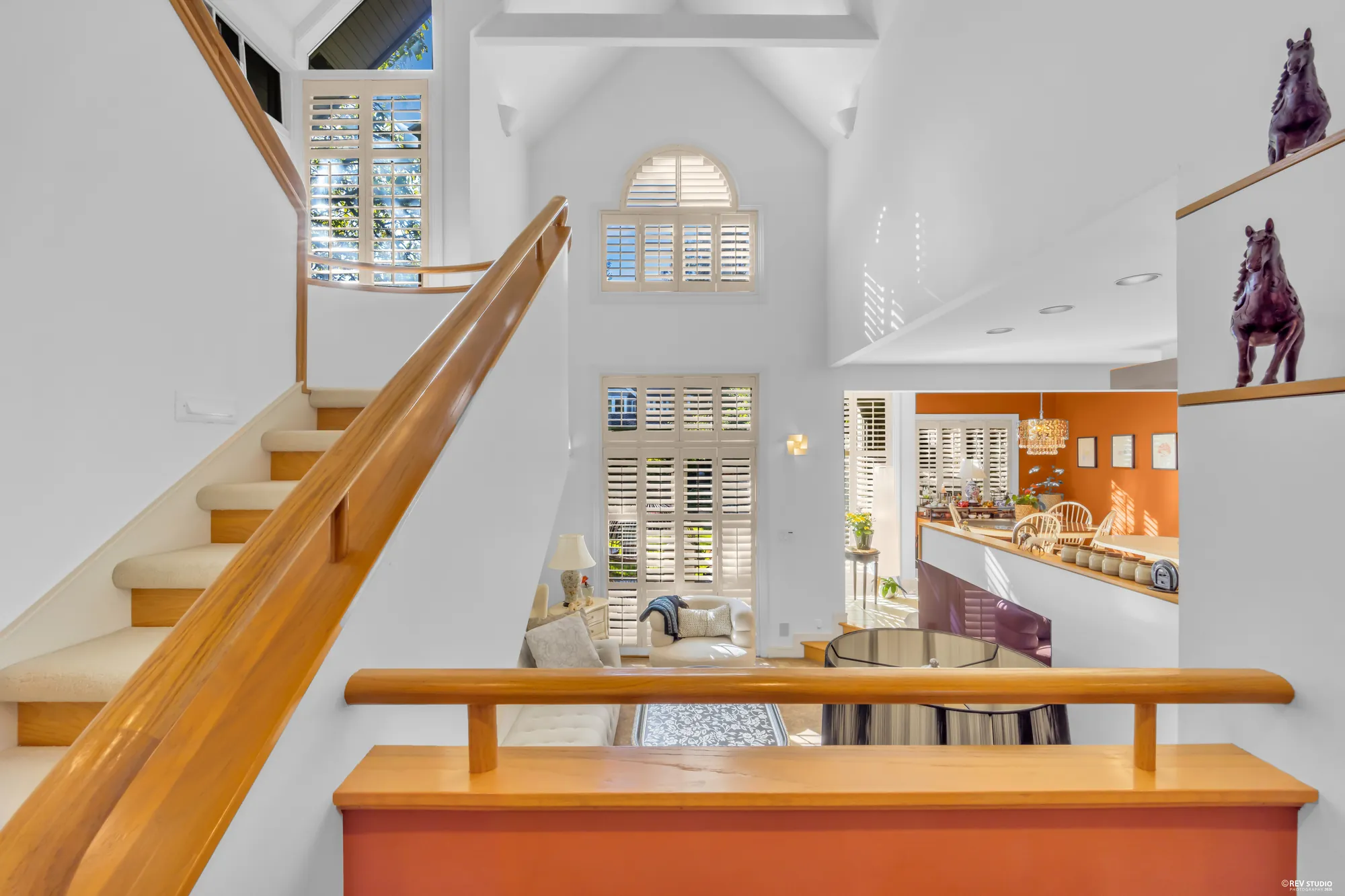 The oak stair rising from the foyer, its rail curving up past tall shuttered windows, the kitchen's terracotta soffit through the opening on the right