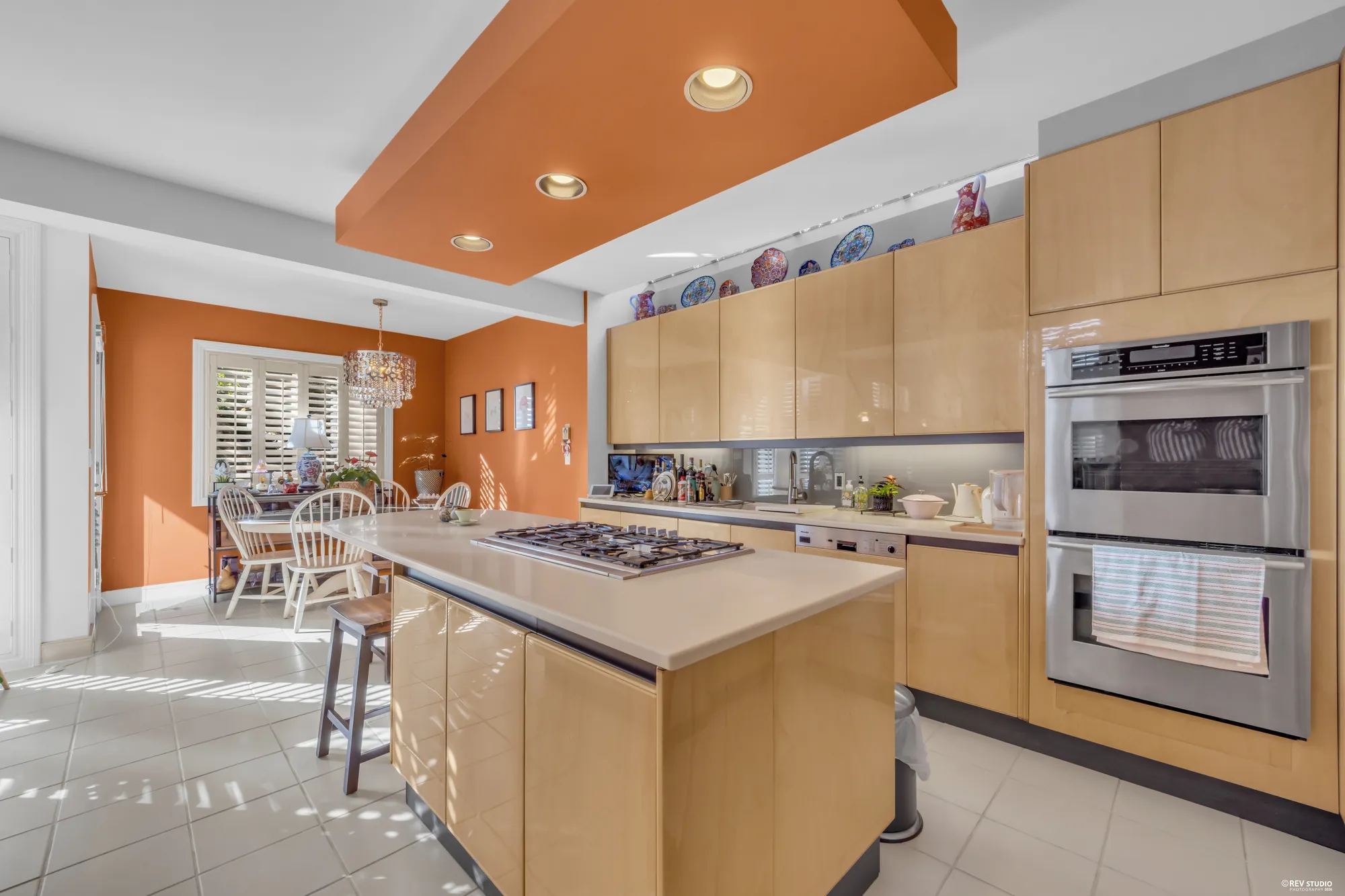The kitchen: pale flat-front cabinets under a terracotta soffit, an island with a gas cooktop and bar stools, double wall ovens, the nook and its shuttered window beyond
